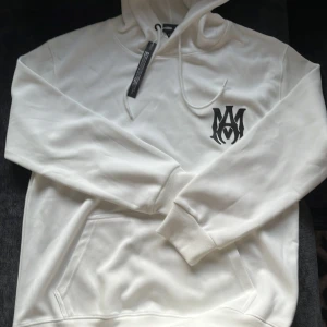 Vit hoodie från Amiri med tryck - Snygg vit hoodie från Amiri i bomull med svart logga på bröstet och stort AMIRI-tryck på ryggen. Klassisk känguruficka, huva med snörning och ribbade muddar. Perfekt för dig som gillar streetwear och vill sticka ut med stilren design.