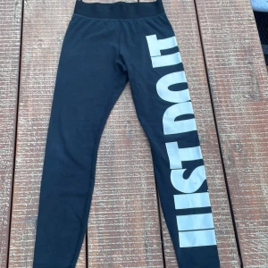 Nike leggings med stor JUST DO IT-text - Svarta leggings från Nike med stor vit 'JUST DO IT.'-text längs ena benet. Tight passform och elastisk midja. Perfekta för träning eller chill. Tillverkade i mjukt och stretchigt material för maximal komfort.