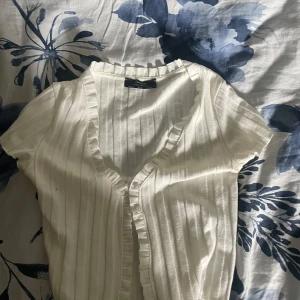 Vit ribbad blus från Bershka XS - Supersöt vit ribbad blus/topp från Bershka i storlek XS. Den har korta ärmar, v-ringning med volangkant och knappar på framsidan. Använd Max 2 gånger ser helt ny ut 