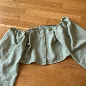 Ljusgrön offshoulder blus med knappar - Trendig ljusgrön blus med offshoulder-modell och långa puffiga ärmar. Toppen har en snygg knapprad framtill och är gjord i ett strukturerat tyg som känns både luftigt och coolt. Perfekt för dig som gillar en romantisk och avslappnad stil.
