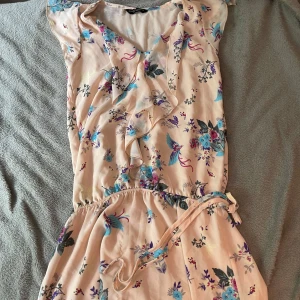 Blommig kortklänning med volang - Supersöt kortklänning i ljusbeige med blommigt mönster i blått, lila och rosa. Klänningen har volangdetaljer framtill, v-ringning och ett knytband i midjan. Perfekt för sommarens alla tillfällen och ger en romantisk vibe.