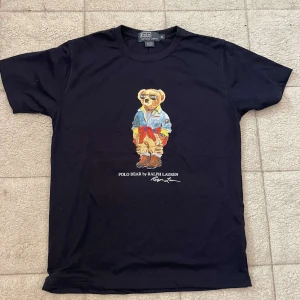 Mörkblå Polo Bear t-shirt från Ralph Lauren - Mörkblå t-shirt från Polo Ralph Lauren med ikoniskt Polo Bear-tryck på bröstet. T-shirten är i bomull, har rund hals och korta ärmar. Snygg och klassisk modell med färgglad björn i streetwear-stil framtill. Perfekt för dig som gillar statement-plagg.