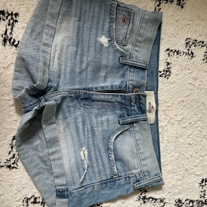 Ljusblå jeansshorts från Hollister - Snygga ljusblå low waisted jeansshorts från Hollister med slitna detaljer och klassisk femficksdesign. Shortsen har uppvikta benslut, dragkedja och knapp framtill samt dekorativa sömmar på bakfickorna. Perfekta för varma dagar och en avslappnad stil. W26🥰