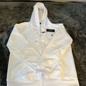 Vit hoodie från Polo Ralph Lauren - Snygg vit hoodie från Polo Ralph Lauren med dragkedja och klassisk huva. Tröjan har broderad logga på bröstet, ribbade muddar och fickor framtill. Perfekt för en clean och stilren look. Materialet är mjuk bomull som känns skönt mot huden.