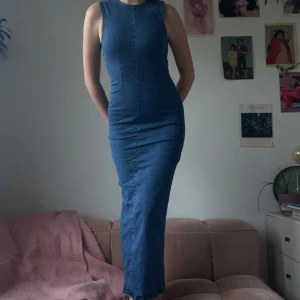 Blå jeansklänning - Säljer en lång, ärmlös jeansklänning från Gina Tricot i klassisk blå denim. Klänningen har rund halsringning, figurnära passform och stängs med knapp i nacken. Materialet är mjukt och stretchigt med 98% bomull och 2% elastan.