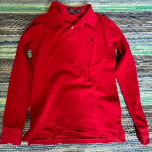 Röd långärmad pikétröja Polo Ralph Lauren - Röd långärmad pikétröja från Polo Ralph Lauren med klassisk krage och två knappar framtill. Tröjan är i bra skick uta några hål eller liknande. Bara höra av sig vid frågor.