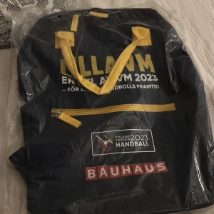 Handbollsväska Polen Sverige 2023 - Mörkblå sportig väska med gula handtag och dragkedja, tryck för Handbolls-VM 2023 Polen/Sverige och Bauhaus-logga. Perfekt för dig som gillar handboll och vill ha en unik väska med coolt tryck och praktiskt fack framtill. Helt oanvänd aldrig upp pakad.