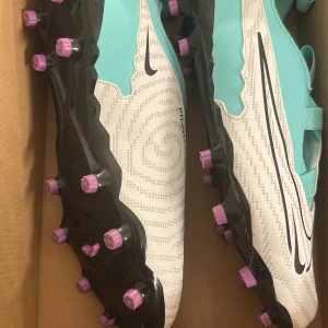 Nike Phantom GX fotbollsskor  - Nike Phantom GX Academy FG/MG fotbollsskor i fräsch turkos och vit med svarta detaljer och lila dobbar. 