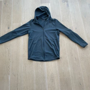 Svart träningsjacka från Under Armour - Sportig svart vindjacka från Under Armour med huva och hel dragkedja. Jackan är tillverkad i ett mjukt syntetmaterial och har två fickor framtill. Perfekt för träning eller när du vill ha en clean look. Diskret logga på bröstet.