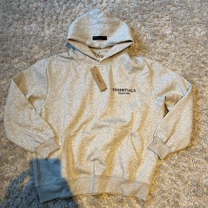 Grå Essentials Fear of God hoodie - Snygg grå hoodie från Essentials Fear of God med svart logga på bröstet och ryggen. Klassisk passform med huva och känguruficka. Mjuk bomullsblandning som är perfekt för chill dagar.                                             Om du skulle ha nån fråga så e de bara att skriva.