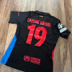 FC Barcelona Lamine Yamal tröja Nike - Snygg FC Barcelona fotbollströja med Lamine Yamal och nummer 19 på ryggen. Svart bas med röda och blå detaljer, officiella loggor från Nike, Spotify och Champions League. Tillverkad i lätt, ventilerande Dri-FIT material. Perfekt för dig som älskar Barca och vill sticka ut på planen.