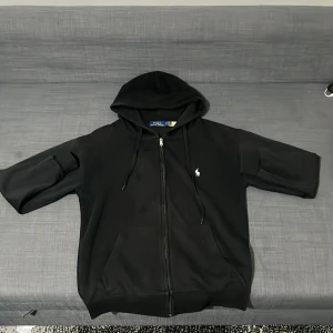 Svart hoodie från Polo Ralph Lauren - Svart hoodie från Polo Ralph Lauren med dragkedja och huva. Klassisk design med vit broderad logga på bröstet, känguruficka och snörning i huvan. Skön passform och perfekt för chill dagar eller när du vill hålla det stilrent. inga fläckar då det är bara vatten droppar.