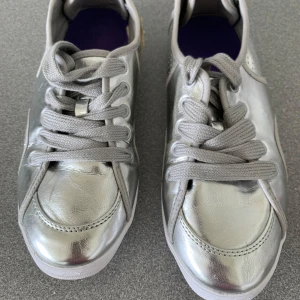 Silvriga sneakers med snörning - Snygga sneakers i metallic-silver med grå snörning och vit sula. Skorna har rund tå och perforerade detaljer på sidorna för extra stil. Perfekta för dig som vill sticka ut med en futuristisk vibe. Ovandelen är i syntetmaterial med glansig finish.