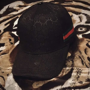 Svart Gucci keps  - Snygg svart keps från Gucci med diskret GG-monogram över hela kepsen och klassiska gröna och röda ränder på sidan. Kepsen har böjd skärm och justerbar rem baktill. Perfekt för dig som vill ha en lyxig och stilren accessoar.