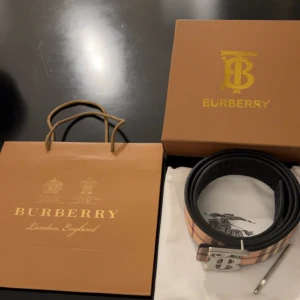 Burberry rutigt skärp med B-spänne - Snyggt Burberry-bälte i klassiskt rutigt mönster med beige, svart och röda toner. Bältet har ett stort silverfärgat B-spänne och är gjort i exklusivt skinn. Perfekt accessoar för att lyfta din outfit med en lyxig touch och ikonisk design. Pris kan diskuteras vid snabb affär. Det är bara att skriva om ni har fler frågor om den.