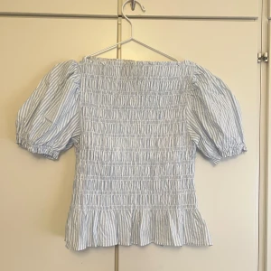 Smockad blus  - Trendig blus i ljusblått och vitt med smockad kropp, puffiga korta ärmar och volang nertill. Tunt bomullstyg med diskreta ränder ger en fräsch sommarkänsla. Priset går att diskuteras :)
