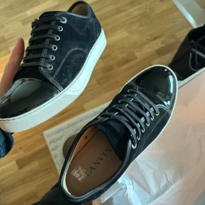 Svarta sneakers från Lanvin med lacktå - Snygga svarta sneakers från Lanvin med ovandel i mocka och en glansig lackad tå. Skorna är köpta på NK för ca 2 veckor sedan. Dem är för små för mig så har fått skavsår. Kvitto finns till, alla tillbehör medföljer och skorna är inpricip nya. 9.5/10 i skick. Size uk 8 vilket motsvarar 42/43