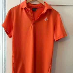 Orange pikétröja från Cross Sportswear - Snygg orange pikétröja från Cross Sportswear med klassisk krage och korta ärmar. Tröjan har knappar vid halsen och en vit broderad logga på bröstet. Perfekt för en sportig och fräsch look under sommaren.