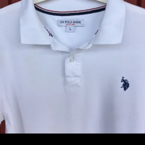 Vit pikétröja från U.S. Polo Assn. - Klassisk vit pikétröja från U.S. Polo Assn. med broderad logga på bröstet. Tröjan har krage, knäppning med två knappar och korta ärmar. Perfekt för en clean och sportig stil.