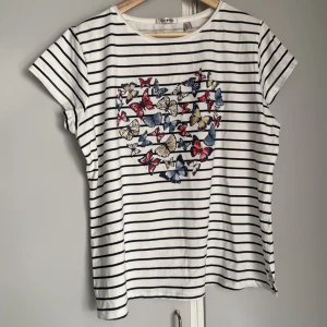 Randig t-shirt med fjärilar från Punt o Roma - Vit och svart randig t-shirt från Punt o Roma med korta ärmar. Framsidan har ett stort tryck av färgglada fjärilar i rött, blått och beige med strass. Storlek Large