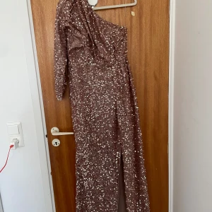 Glittrig one shoulder långklänning - Säljer en glittrig långklänning i brunbeige ton med paljetter över hela klänningen. Den har en ärm och är oneshoulder, samt en snygg slits framtill. Perfekt för dig som vill sticka ut och glänsa på festen!