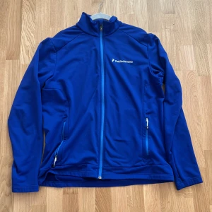 Blå vindjacka från Peak Performance - Snygg blå vindjacka från Peak Performance med hel dragkedja och två fickor med dragkedja. Jackan har hög krage och är perfekt för dig som gillar en sportig stil. Materialet är mjukt och smidigt, vilket gör den skön att bära under kyliga dagar.