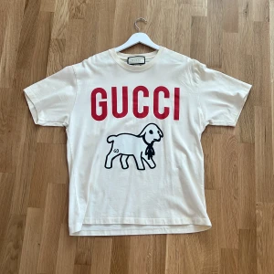 Vit GUCCI t-shirt med lammtryck - fick i födelsedagspresent och inte andvänt.ps den är äkta.Vit t-shirt från GUCCI med stor röd logga och ett svartvitt lammtryck framtill. Klassisk rund hals och korta ärmar. Tillverkad i mjuk bomull och perfekt för sommaren. 