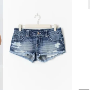 Blå slitna jeansshorts med fransar - Snygga blå jeansshorts med slitna detaljer och fransig kant. Klassisk femficksmodell med låg midja och dragkedja. Perfekta för varma sommardagar och en avslappnad streetstyle-look.