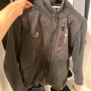 Svart Sail Racing hoodiejacka med dragkedja - Svart hoodiejacka från Sail Racing med hel dragkedja, bröstficka med dragkedja och broderad logga på bröstet. Jackan har huva med tryck och är tillverkad i mjukt material, perfekt för chill dagar. Snygga detaljer och sportig look.