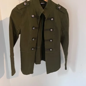 Olivgrön rock från Gina Tricot, stl 36 - Snygg olivgrön rock från Gina Tricot i militärinspirerad stil med dekorativa knappar framtill och axelklaffar. Jackan har lång ärm och markerad midja, perfekt för dig som vill sticka ut med en cool look.