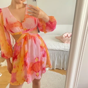 Färgglad kortklänning med cut-outs - Superfin kortklänning i rosa, orange och gul med akvarellmönster. Klänningen har v-ringning, långa puffärmar och snygga cut-outs i midjan. Perfekt för sommaren med sitt luftiga material och lekfulla look.