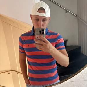 Randig pikétröja från Ralph Lauren - Säljer en snygg pikétröja från Polo Ralph Lauren i custom slim fit. Tröjan har breda blå och röda ränder, klassisk krage och korta ärmar. Perfekt för dig som gillar en sportig och stilren look. Ikoniska loggan broderad på bröstet. 