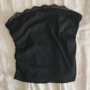 Svart bandeau tubtopp med spets - Säljer en svart bandeau tubtopp med ribbad struktur och spetskant upptill. Toppen har en liten rosett framtill och är helt axelbandslös, perfekt för en stilren och trendig look. Storlek S passar även liten M 