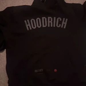 Svart hoodie från Hoodrich - Svart hoodie från Hoodrich med stor logga i grått över bröstet och mindre tryck på fickan. Klassisk huva med snörning och känguruficka framtill. Perfekt för en streetwear-look och chill dagar.