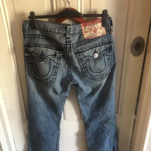 True Religion Billy Super T jeans - Säljer ett par blå True Religion Billy Super T jeans med slitna detaljer och hål på benen. Jeansen har kontrastsömmar, stora fickor med lock och silverfärgade knappar. Klassisk femficksmodell i bomull med låg midja och rak passform.