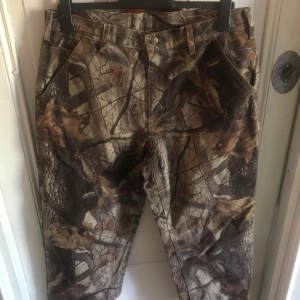 Carhartt workpants x realtree - Säljer ett par Carhartt workpants med fett camouflage-mönster i bruna och gröna toner. Byxorna har flera stora fickor, robust bomullstyg och klassisk Carhartt-patch bak. Perfekta för dig som gillar streetwear eller outdoor-vibe.