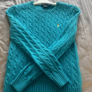 Turkos kabelstickad tröja Ralph Lauren - Säljer en turkos kabelstickad tröja från Ralph Lauren med klassisk gul broderad logga på bröstet. Tröjan har rund halsringning, långa ärmar och ribbade muddar. Perfekt för dig som vill ha en färgstark och tidlös look.