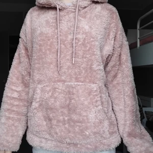 Rosa fluffig hoodie från H&M - Supermjuk och oversized hoodie från H&M i en ljusrosa ton. Tröjan har huva med snörning, stor magficka och är tillverkad i fluffigt syntetmaterial. Perfekt för kalla dagar när du vill mysa till det extra.