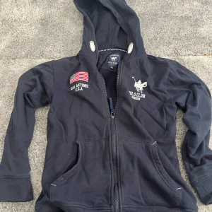 Mörkblå hoodie från Vinson Polo Club - Mörkblå hoodie med dragkedja från Vinson Polo Club. Tröjan har huva, broderad amerikansk flagga och logga på bröstet samt fickor framtill. Perfekt för en sportig och avslappnad stil. Tillverkad i mjuk bomull för extra komfort.
