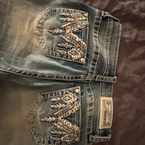  Low waist Grace blå jeans med broderade fickor - Snygga blå jeans från Grace med broderade bakfickor och paljettdetaljer. Jeansen har bootcut-modell och kontrastsömmar, samt en tvättad look med ljusare partier. Perfekta för dig som gillar unika detaljer och en cool stil. Nypris 2000 säljer för 1200