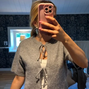 Grå kofta med snörning från Gina Tricot - Supermjuk och fluffig grå kofta från Gina Tricot i ullblandning. Kortärmad modell med snygg snörning framtill och ribbade kanter. Perfekt att slänga över ett linne för en chill och trendig look.