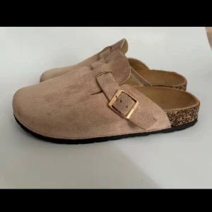 Beige mockasandaler med spänne - Snygga slip-in sandaler liknande Birkenstock i beige mocka med guldfärgat spänne på sidan. 