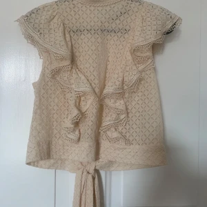 Beige spetsblus med volang och knyt - Supersöt beige blus i spets med volangdetaljer längs axlar och fram, samt knytband nedtill. Blusen har genombrutet mönster, dekorativa knappar i nacken och romantisk känsla. Perfekt för dig som gillar detaljer och en feminin stil. Köparen betalar frakten 