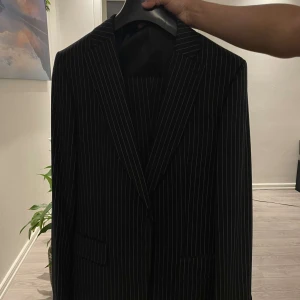 Svart randig kostym från Next Tailoring - Snygg svart kostym från Next Tailoring med tunna vita rände kostym byxan följs med😁. Säljer den för att den inte används länger. Har haft den på mlg engång. Helt ny. Pris är inte huggen på sten😊