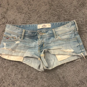 Ljusblå jeansshorts från Hollister - Snygga ljusblå jeansshorts från Hollister med slitna detaljer och råa kanter. Klassisk femficksmodell med knapp och dragkedja framtill, samt broderad logga på fickan. Perfekta för varma dagar och en avslappnad stil.