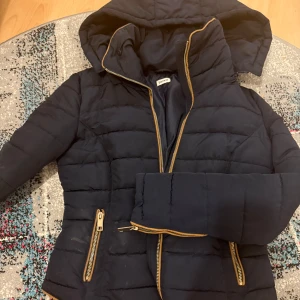 Mörkblå pufferjacka från Pimkie - Snygg mörkblå pufferjacka från Pimkie med guldiga dragkedjor och huva. Jackan har quiltad design, långa ärmar och figurnära passform. Perfekt för kyliga dagar när du vill hålla stilen enkel men ändå trendig. Jackan har använts en vinter och inget mer. Kommer naturligtvis kemtvätts den innan den skickas. Ord pris 699kr, pris kan diskuteras.