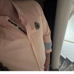 Randig skjorta från Morris, M - Snygg ljusorange och vitrandig skjorta från Morris med broderad patch på bröstet. Skjortan har klassisk krage, knäppning framtill och långa ärmar med kontrastfärg på insidan av manschetten. Tillverkad i bomull för en skön känsla.
