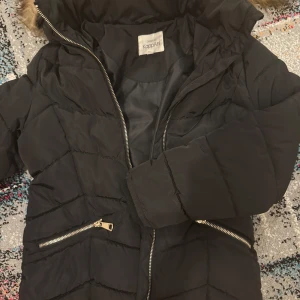 Svart pufferjacka från KappAhl - Svart pufferjacka från KappAhl med snygga guldiga dragkedjor och huva med fejkpäls. Jackan har quiltad design och långa ärmar, perfekt för kalla dagar. Praktiska fickor med dragkedja och reflekterande detalj på ryggen. Jackan användes en vinter. Ord pris 499kr.