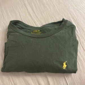 Grön t-shirt från Polo Ralph Lauren - Snygg grön t-shirt från Polo Ralph Lauren med klassisk gul broderad logga på bröstet. T-shirten har rund halsringning och är tillverkad i mjuk bomull, perfekt för en chill och stilren look.