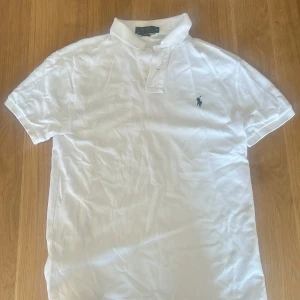Vit pikétröja från Polo Ralph Lauren - Klassisk vit pikétröja från Polo Ralph Lauren med blå broderad logga på bröstet. Tröjan har korta ärmar, krage och knäppning framtill. Slim fit-modell i mjuk bomull som passar perfekt till jeans eller shorts.